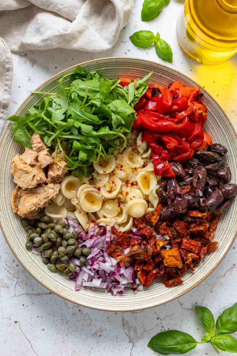 Preparing tuna pasta salad recipe with sweet roasted red peppers, sun-dried tomatoes, Kalamata olives, capers and arugula leaves