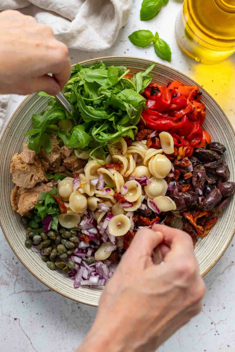 Preparing tuna pasta salad recipe with sweet roasted red peppers, sun-dried tomatoes, Kalamata olives, capers and arugula leaves