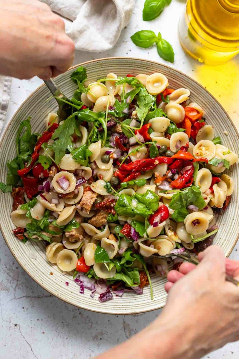 Preparing tuna pasta salad recipe with sweet roasted red peppers, sun-dried tomatoes, Kalamata olives, capers and arugula leaves
