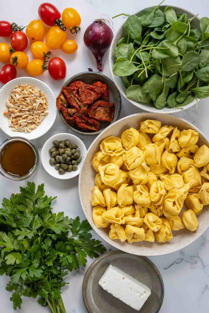 Nudelsalat Zutaten: Feta, Spinat, gefüllten Nudelteigtaschen, Petersielle, Kapern, Tomaten, Mandeln, rote Zwiebel und Dressing
